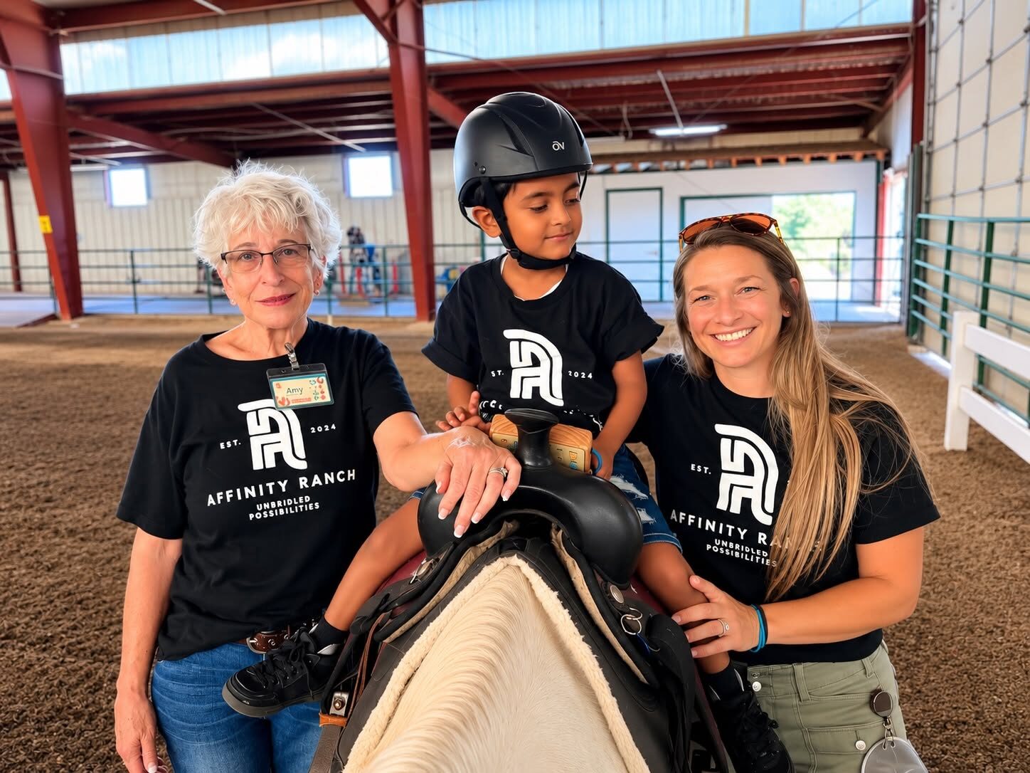 Two Affinity Ranch volunteers in black Unbridled Possibilities t-shirts steadying a young rider in a riding helmet seated in the saddle during an adaptive horsemanship session in the ranch's indoor arena