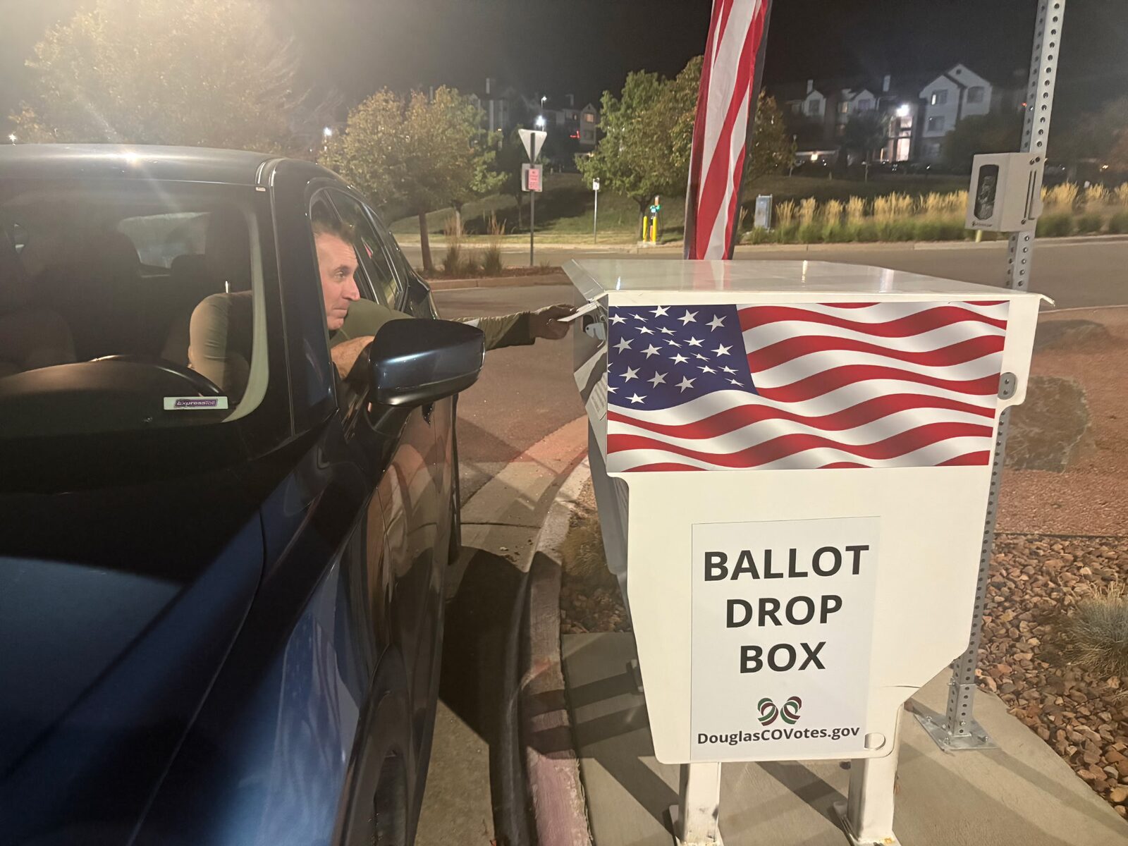 Tim Dietz leaning out the window of his car at night to drop his ballot into a Douglas County ballot drop box decorated with an American flag and marked DouglasCOVotes.gov