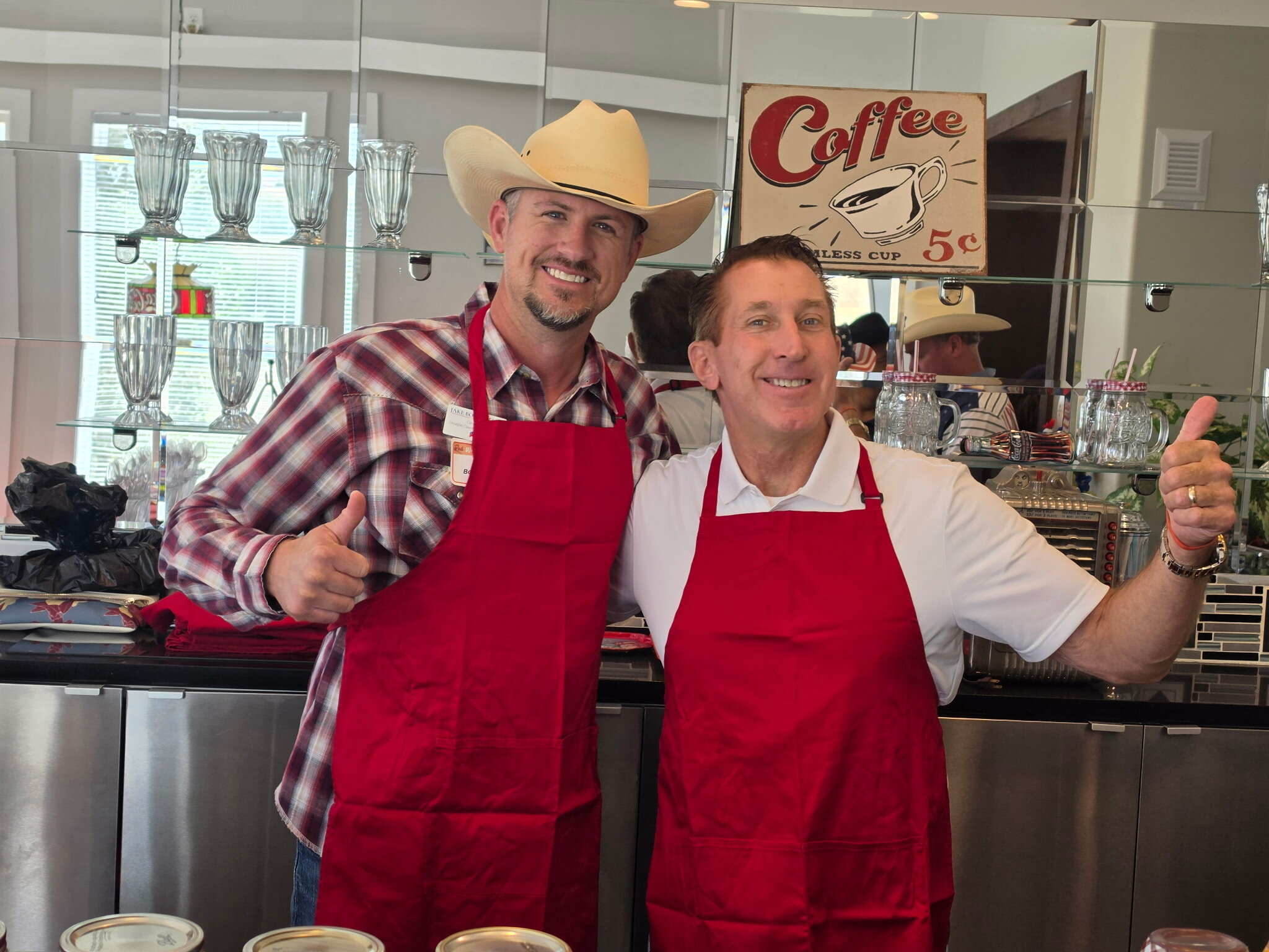 Jake Bockenfeld, left, and Tim Dietz, right, in red aprons giving thumbs up in front of a retro Coffee sign while serving ice cream at the Douglas County Republican Women luncheon and fundraiser