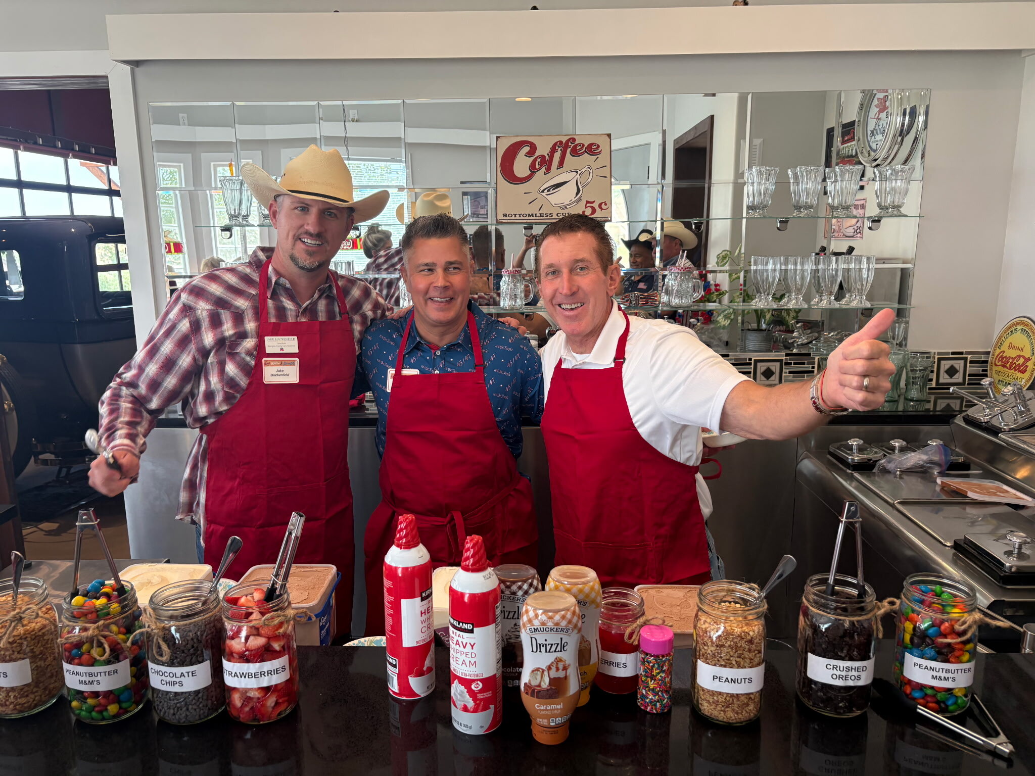Jake Bockenfeld in a cowboy hat, Sheriff Darren Weekly, and Tim Dietz in red aprons behind a vintage soda-fountain counter lined with sundae toppings at the Douglas County Republican Women luncheon and fundraiser
