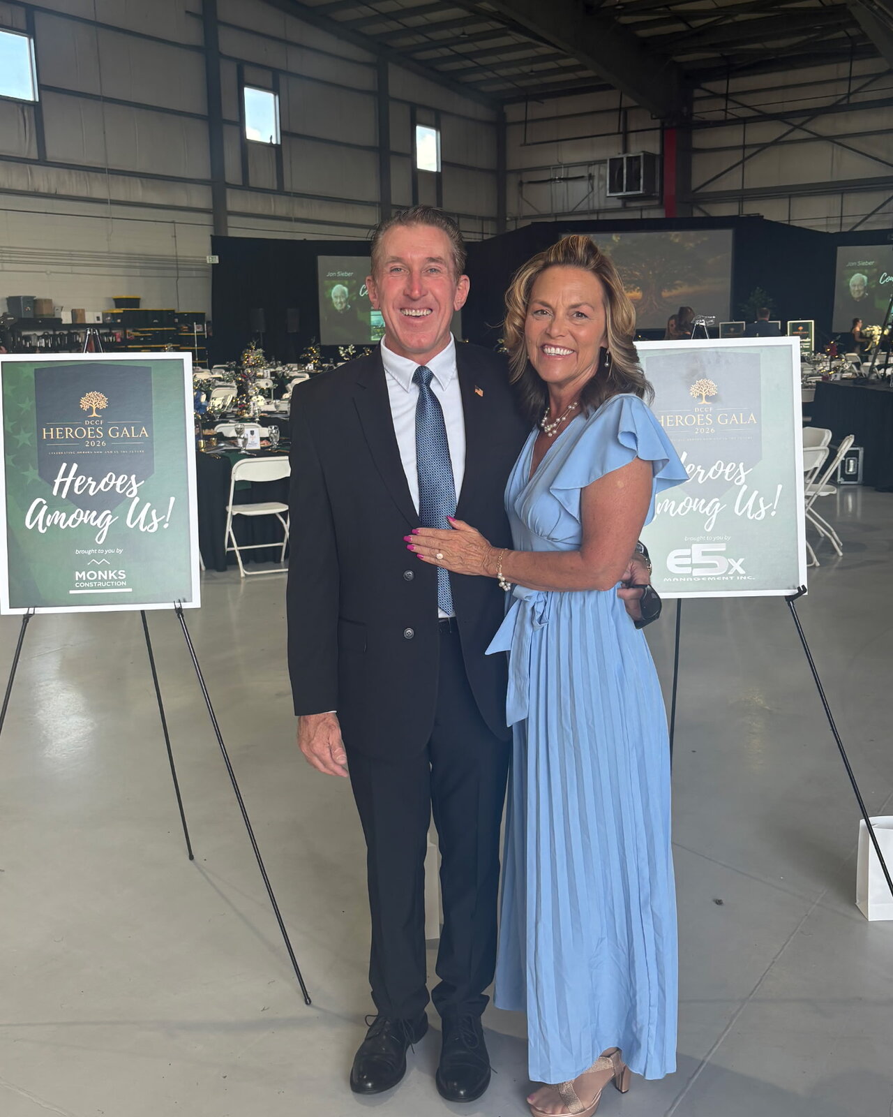 Tim Dietz in a dark suit and his wife JoAnna in a light blue gown inside the hangar at the 2026 Douglas County Heroes Gala, between two easel signs reading Heroes Gala 2026 — Heroes Among Us
