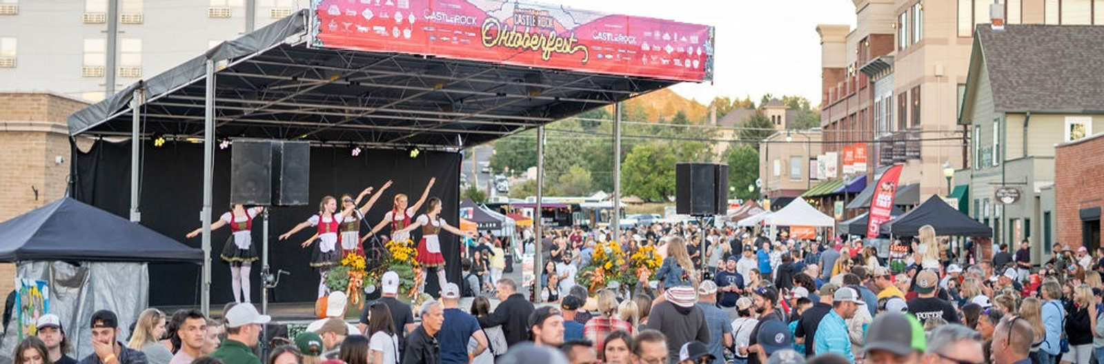 Crowd filling Wilcox Square at Castle Rock Oktoberfest as dancers in dirndls perform on the main stage under the Oktoberfest banner, with downtown storefronts and food trucks in the background