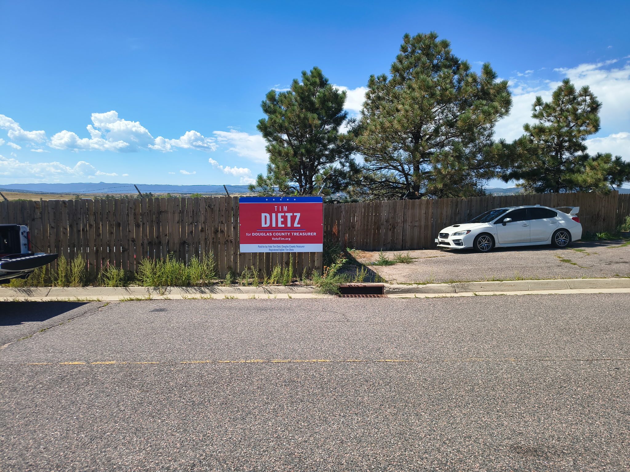 A red, white, and blue Tim Dietz for Douglas County Treasurer campaign sign mounted on a wooden fence in Sedalia by O'Brien's Cafe, with pines and the Front Range in the distance